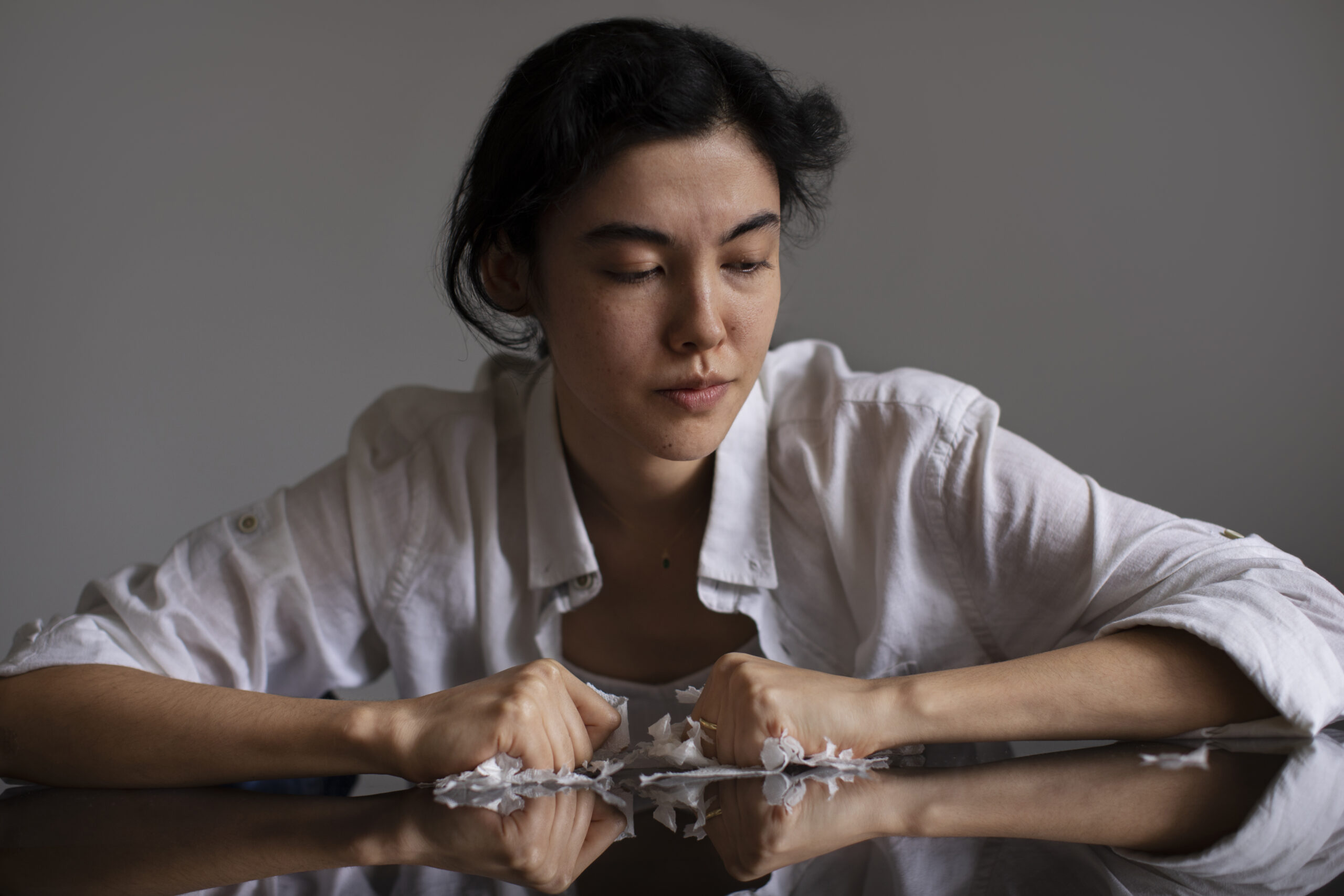 A woman sits at a table looking down with a tense expression, her hands clenched among crumpled pieces of paper, suggesting feelings of frustration or anxiety.