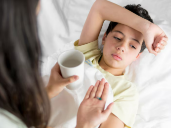 A young boy lying in bed looking unwell while an adult hands him a cup of water and a pill
