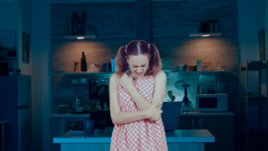 A young girl in a checkered dress standing in a dimly lit kitchen, holding her arm and appearing distressed or in pain