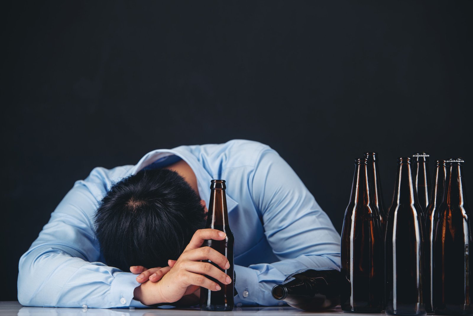 A man sitting at a table with his head down surrounded by empty beer bottles, showing signs of exhaustion or alcohol abuse.