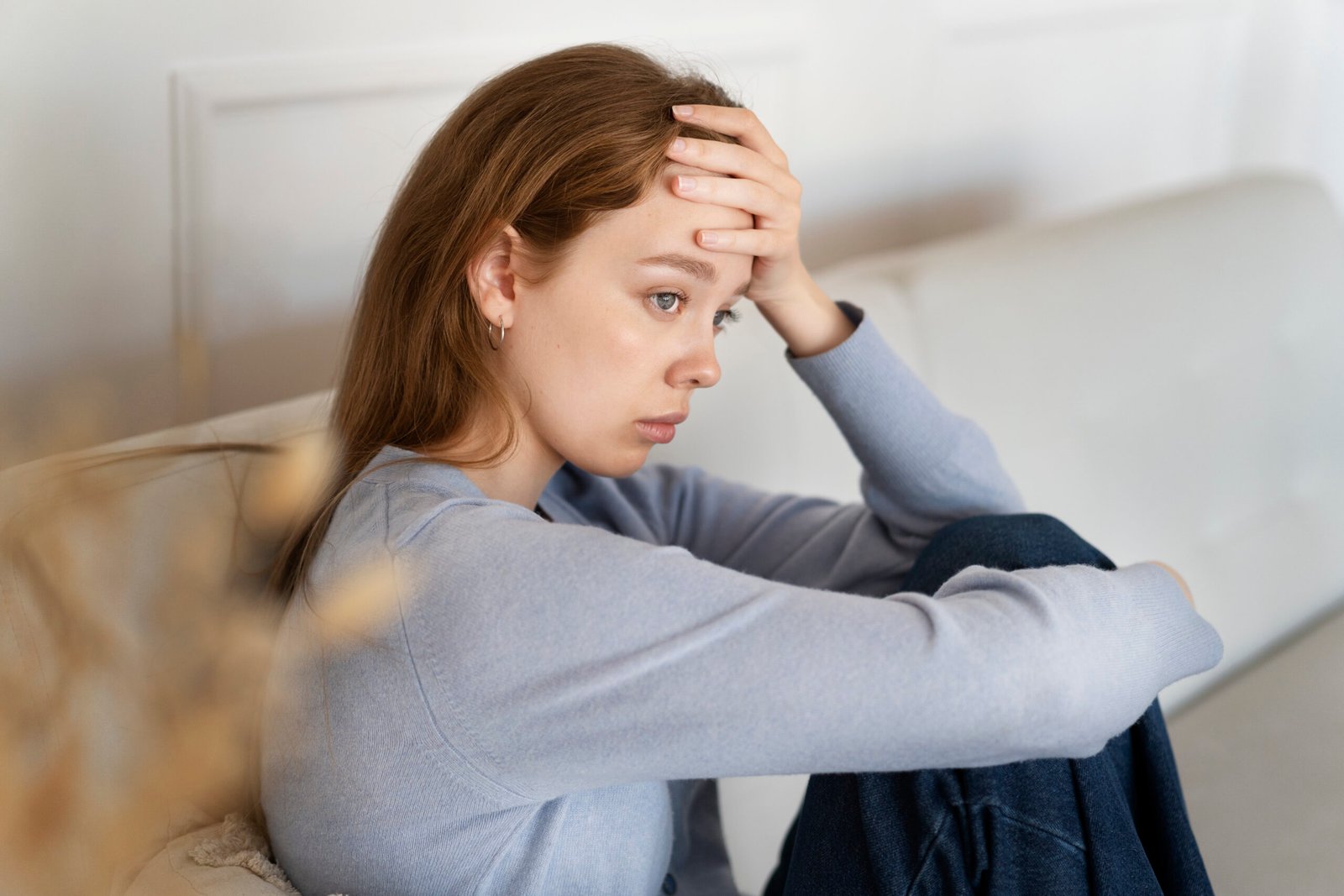 A young woman sitting on a couch with her hand on her forehead, appearing stressed or deep in thought