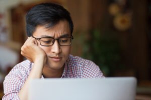 A man wearing glasses looks concerned while staring at a laptop screen, resting his head on his hand