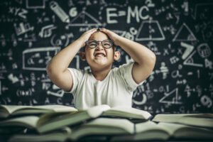 A frustrated young boy wearing glasses sits at a desk full of open books, holding his head with both hands, with mathematical formulas and drawings on the blackboard behind him — representing the challenges children with ADHD may face in learning.
