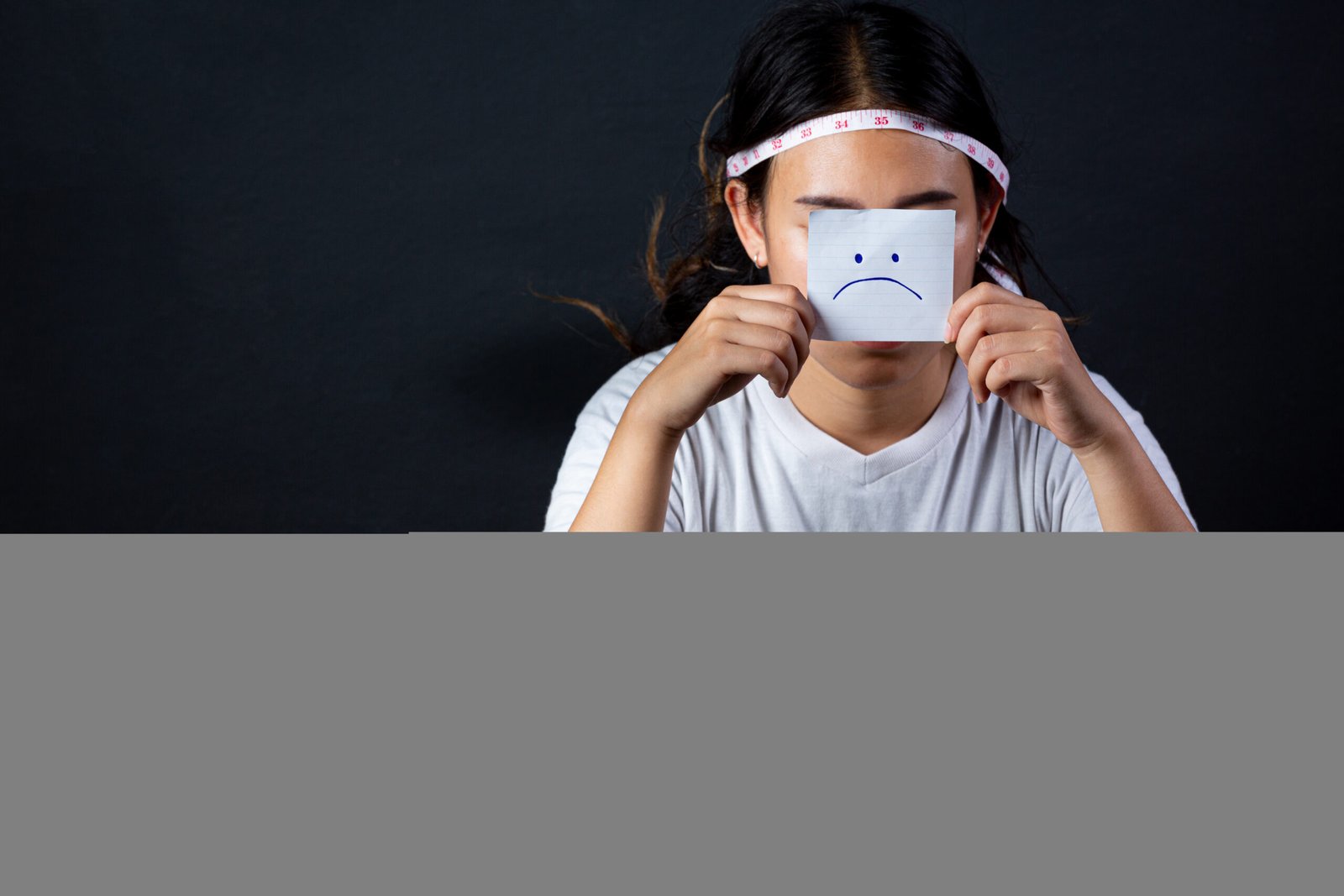 Một người phụ nữ đội thước dây quanh đầu, cầm tờ giấy có hình mặt buồn che mặt, ngồi trước đĩa rau xanh nhỏ trên bàn trong bối cảnh nền tối.