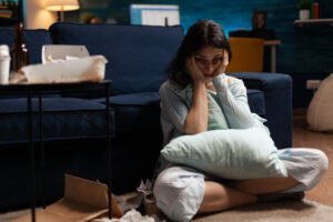 A woman sitting on the floor in a messy living room, hugging a pillow and looking exhausted and overwhelmed, with takeout boxes and tissues scattered around