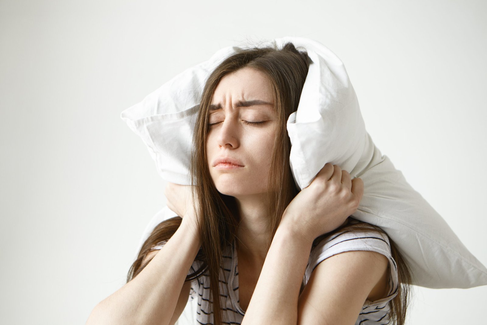 Young woman covering her ears with a pillow, showing frustration and difficulty sleeping due to noise or stress