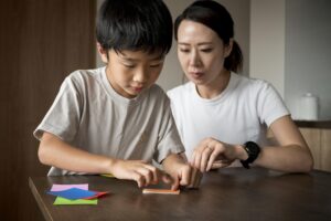 A mother helping her young son with a learning activity at a table, using colorful paper cards to support his cognitive and motor development.