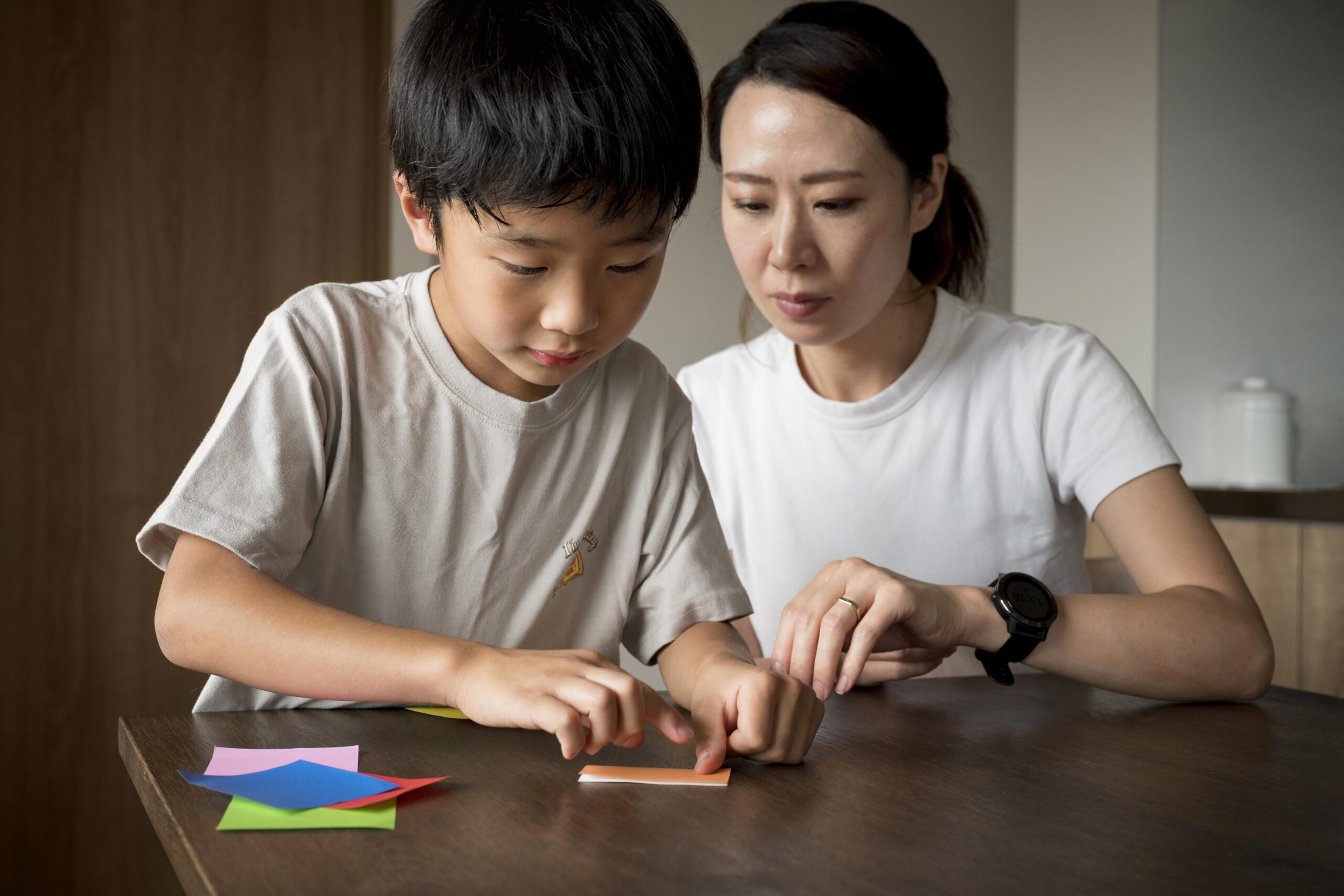A mother helping her young son with a learning activity at a table, using colorful paper cards to support his cognitive and motor development.