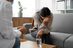 A woman sitting on a couch appears distressed, wiping her eyes while pointing toward a glass of water, as a therapist sits nearby listening attentively.