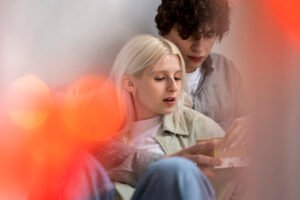 A couple sitting closely together, with the man embracing the woman from behind as they look at a small gift box