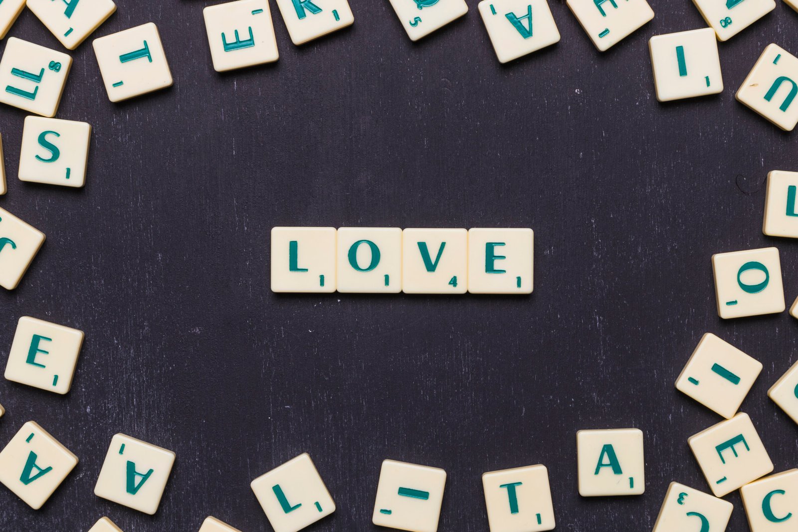 A group of scattered Scrabble tiles surrounds four centered tiles that spell the word “LOVE” on a dark background
