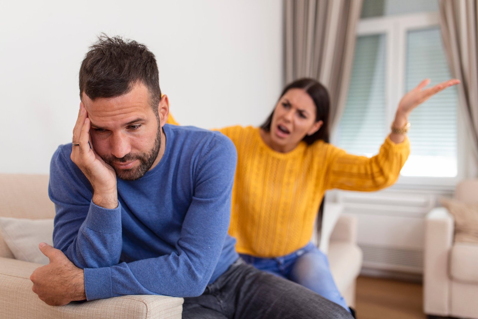 A woman in a yellow sweater appears to be angrily gesturing while a man sitting in front of her looks upset and withdrawn, resting his head on his hand in a living room setting.