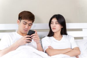 A woman sitting in bed with her arms crossed, looking annoyed at a man next to her who is focused on his phone and not paying attention to her.